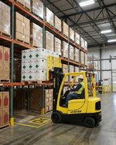 Person operating a yellow Hyster forklift in a warehouse with stacked boxes.